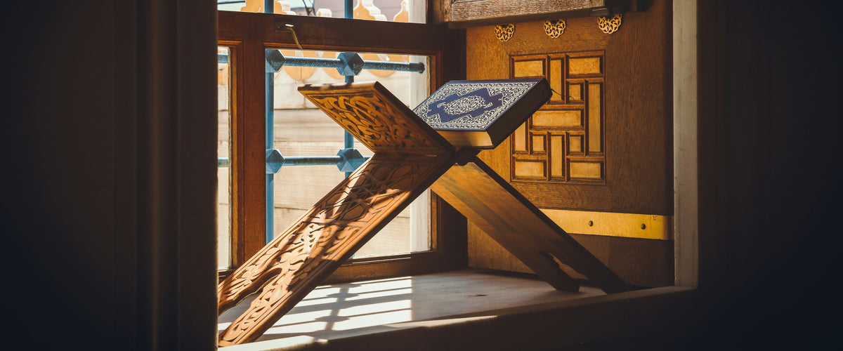 An open Qur'an resting on a carved wooden stand in a sunlit mashrabiya window
